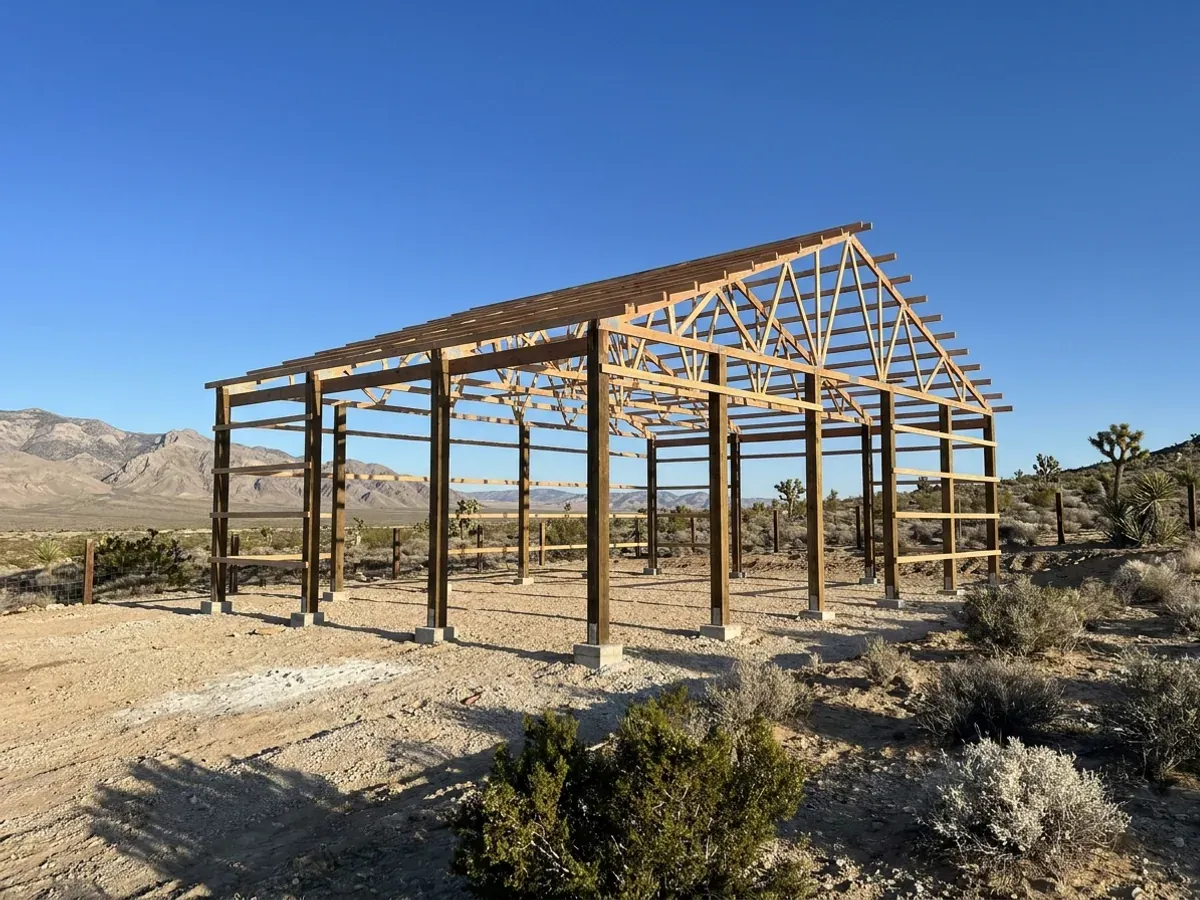 A post-frame home under construction on a desert lot, showing laminated timber columns on concrete piers and wide-span roof trusses.
